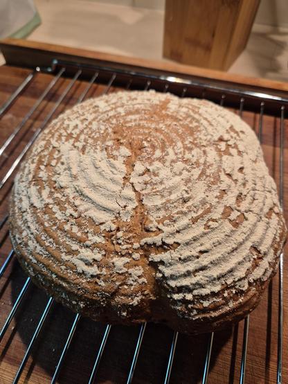 A loaf of freshly baked bread sits on a cooling rack, set against a neutral background. The bread appears golden brown and crusty, emanating a warm, inviting aroma. The composition of the image is simple yet elegant, showcasing the artistry of baking. The dominant colors of brown and black create a cozy and rustic atmosphere. The bread is the focal point of the image, highlighting its texture and shape.