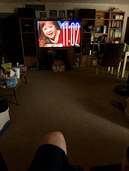 A dimly lit living room is shown from the perspective of someone seated on a chair or couch, with their knees visible at the bottom of the image. In the center, there is a large TV displaying the time “11:02” in white numbers on a blue and red background. The TV is flanked by wooden shelves and cabinets filled with books, pictures, and various household items. To the left, a table has snacks, drinks, and tissues, while to the right, a dining table and chair are partially visible. The room appears cozy and lived-in, with warm lighting and carpeted floors.