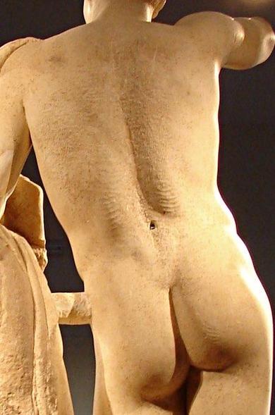 Reverse view of a marble sculpture of Hermes. We see him leaning on a column, his back and buttocks with the legs and head not visible. He's standing on his right leg, the buttock taut, while the left is relaxed.