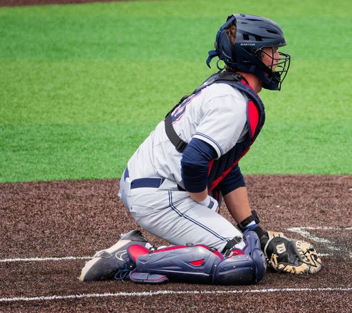 Catcher in baseball crouching behind home plate with grey shoes and shin guards, lighter grey pants and jersey and a blue protective helmet