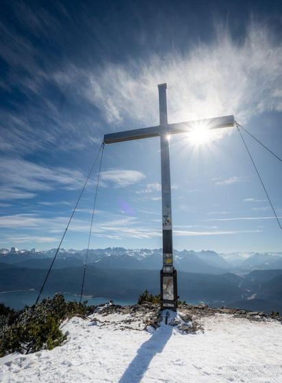 A peaceful scene captured on a snowy mountain, showing a white cross standing tall against a backdrop of grey clouds in the sky. The winter landscape is serene and cold, with a hint of sunlight breaking through. A lone pole is visible nearby, adding to the tranquil atmosphere. The image is devoid of any objects or faces, focusing solely on the beauty of nature and the religious symbol. The dominant colors of white and grey create a sense of calm, while the accent color of deep blue adds a touch…