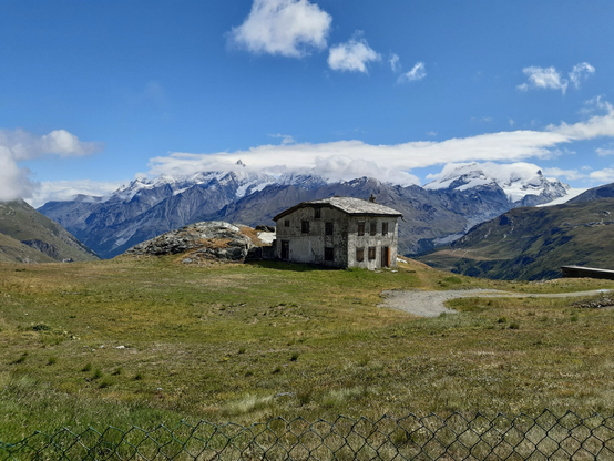 Im Vordergrund eine Wiese mit Steinhüttchen, in der Ferne schneebedeckte Berge