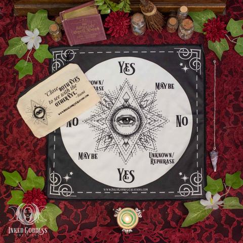 The contents of our Third Eye Pendulum Kit are arrayed on a lace surface. A square black altar cloth is laid flat. It has a large beige circle in the center with third eye imagery line art. Arranged at 8 points around the circle are the words "Yes," "No," "Maybe," and "Unknown/Rephrase." Each word is on the cloth twice, at opposite points on the circle. A brown canvas zipper bag is partially covering the cloth and has a version of the same third eye line art with the text, "Close both eyes to see with the other eye," attributed to Rumi. An amethyst point gemstone pendulum is shown as well as a green tealight for scale. This is an exclusive product from Inked Goddess Creations.