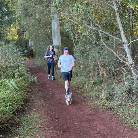 Tow men running on a trail, with the very small terrier type dog on a lead ahead of them, her large furry ears blowing back and tail wagging, obviously enjoying every second