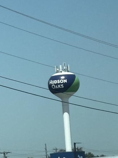 Image of a water tower labeled "HUDSON OAKS," featuring a blue and green design. Power lines run in the foreground against a clear sky.