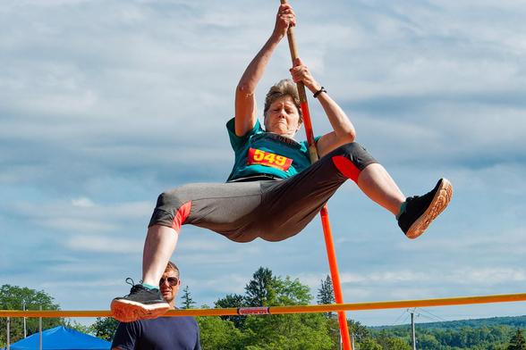 An older woman in a blue shirt with a red bib with yellow 549 holds on to a pole with here hand up in the air with her legs spread out as she flies over the bar while a man looks on from behind;  clouds are in the background of an otherwise blue sky and a few trees can be seen at the bottom