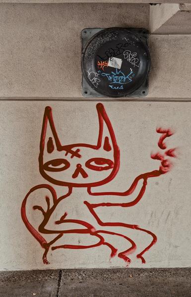 A red line grafiti of a cat with a caret for a nose and two x's on its forhead sitting on a sofa and smoking something with dabs of red paint rising as smoke,  above it is a circular object (bell? air vent?) with a sticker and several grafiti drawn in different colors on it