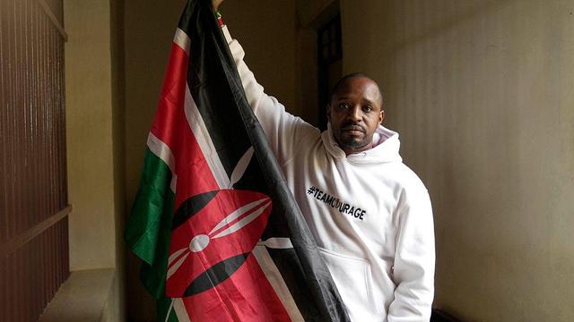 The image shows a man, Boniface Mwangi, standing indoors, holding a flag. The flag is the national flag of Kenya, featuring black, red, green, and white colors with a red shield and a black and white pattern in the center. The man is wearing a white hoodie with the text "#TEAMCOURAGE" printed on it. He is positioned in a corridor with a plain wall and a door visible in the background. The lighting in the image is natural, coming from the left side, casting a soft shadow on the wall behind him.