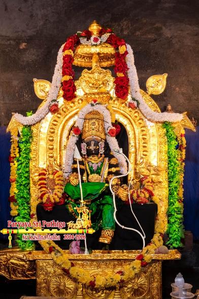Idol of Hindu deity Krishna in green seated with left leg down and right leg folded at the knee and placed over the left knee. Krishna wears gold shoes, gold palm coverings, gold crown, and lots of gold ornaments; holds a silver bow in left hand and a silver arrow in right hand. A string of white flowers around Krishna from his left waist to the right waist, going over the crown.

A gold arch behind Krishna. A thick string of green holy basil leaves and a string of yellow and red flowers along the edges of the arch.