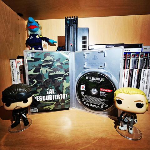 Caja del juego Metal Gear Solid 3: Snake Eater abierta, mostrando el disco en su soporte y el manual de instrucciones a la izquierda. Todo con un diseño interior en tonos oscuros y elementos visuales de la saga.
