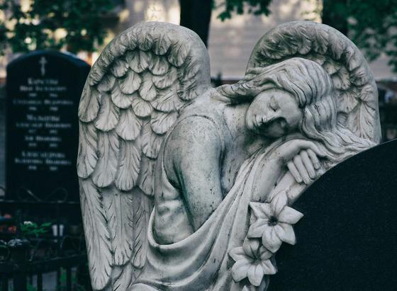victorian cemetary angel statue, female, leading on a tombstone with eyes closed, beautiful and impassive. Winged. The grey pallet and image gives a spooky and or thoughtful impression