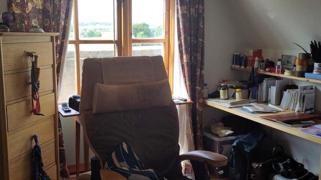 An ergonomic chair sits welcoming in front of two desks and a window with a view of trees receding into the distance