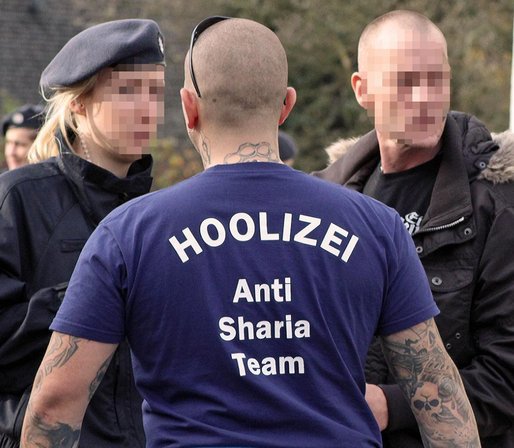 Das Bild zeigt eine Gruppe von Personen, die sich in einem offenen Bereich befinden. Im Vordergrund steht ein Mann mit rasiertem Kopf und tätowierten Armen, der ein tiefblaues T-Shirt trägt, auf dem in großen weißen Buchstaben "HOOLIZEI" steht. Darunter ist in kleineren Buchstaben "Anti Sharia Team" zu lesen. Der Mann trägt eine Sonnenbrille auf dem Kopf und hat mehrere tätowierte Designs auf seinem Nacken und Armen. Neben ihm steht eine Polizistin mit einem dunklen Baret und einem dunklen Mant…