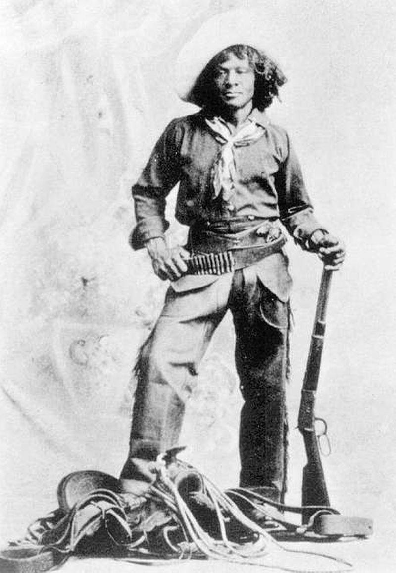 Nat Love stands with a rifle in his hand and wearing a gun belt. A saddle and rope are at his feet.