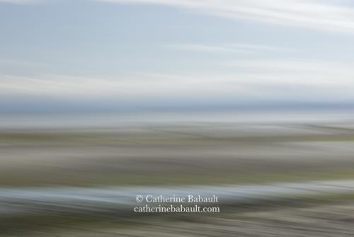 An abstract image of a sandy beach at low tide with some clouds in the blue sky. It looks blurry overall but we can tell where the photo was taken.