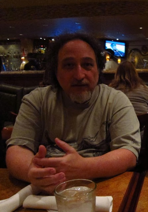 Dave Needle at a restaurant with a drink. He's middle aged, balding, with frizzy hair, a greying goatee, and a quizzical expression. Yes, I had dinner with a few folks and sat across from him.
