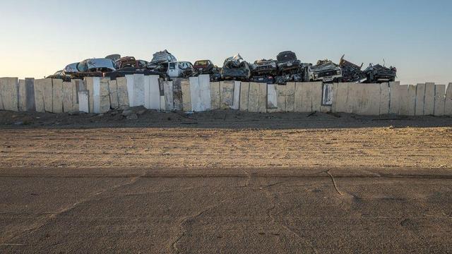 A pile of wrecked cars behind makeshift board fence with sand and paved road in foreground.