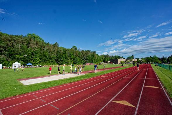 Wide shot along the length of a running track parallel to a long jump,  in the far distance see a high school building, and to the left in the distance a few tents where the event is being run out of.  A line of clouds can be seen in the blue sky to the far right.