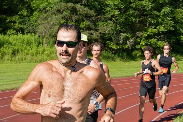 A man with a bare chest and glasses leads a pack of four other runners around the curve of a track with green foliage in the background