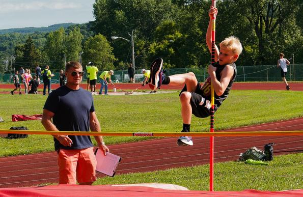 A boy holds on to the pole as he tries to lift himself over the bar with one leg out in front and the other down,  a man in dark glasses, blue t-shirt and orange jeans holds a clipboard and looks on as you can seem people gathered around a long jump pit in the back and a person running on the track in the background