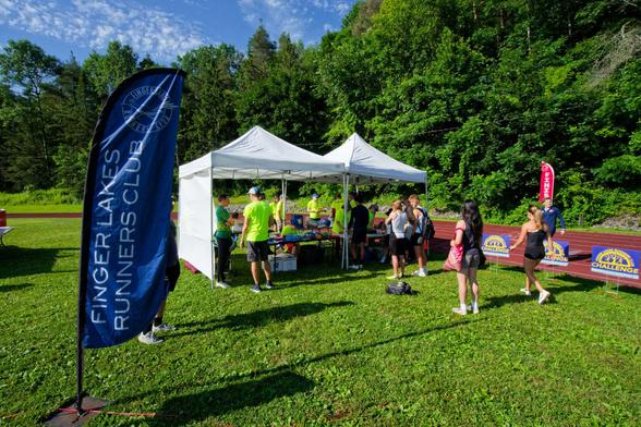 Behind a blue banner that reads FINGER LAKES RUNNERS CLUB find a white tend with two peaks and several volunteers in neon green t-shirts while a few people are waiting outside and a line of blue signs that say FLRC Challenge line the track and a red banner that says FINISH in the background in a sea of green grass and trees