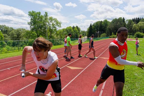 To left a young white man in white shirt and short hair is accelerating forward with a white cylinder in his hand while a black man with even shorter hair and a red to yellow gradient shirt and an arm wrapped up in white breaks to the right.  Way to the right is a young woman in shorts and a splash of pink on her shirt watching while four people stand int he middle of a red track,  behind that there is a short fence and green trees and behind that a dramatic cloudscape of cumulus clouds