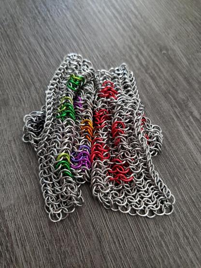 a pile of chainmaille fabric on a gray wood desktop. it is two pieces of fabric in the meant to look like pepperoni pizza when laid out. made with stainless steel and paired with anodized aluminum for the pepperoni slices.