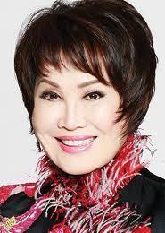 The image features a woman with a short, dark bob haircut, smiling directly at the camera. She is wearing a white, semi-sheer blouse with a large collar and ruffled details, which gives it a sophisticated and elegant appearance. The blouse has a bow detail on the left shoulder. Her makeup is polished, with pink lipstick and defined eyebrows, enhancing her facial features. She is wearing a large, white, diamond-encrusted watch on her left wrist and a matching ring on her left hand. Her nails are…