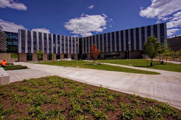 A large building with alternating metal and glass stripes behind a grass and concrete plaza and cumulus clouds behind