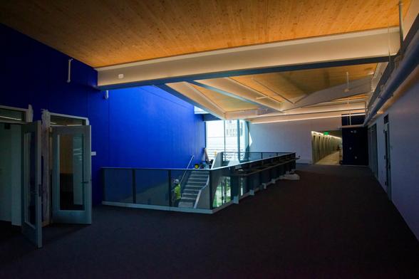 A room with a wood ceiling broken up with various beams and panels and a big blue wall to the far left,  a staricase winding down,  a window to the bright outdoors,  and a corridor leading off to the far right