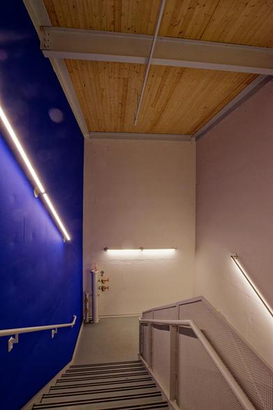 Stairwell with a blue wall to the left and white walls to the rear and white with a pipe for firefighers on the next lading.  The ceiling is made of natural color wood boards with a white metal beam running from left to right and a sprinkler line running from front to back