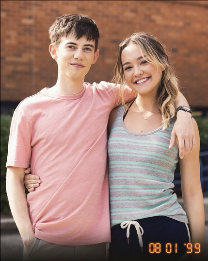 Griffin Gluck and Sadie Stanley, two young white actors, in character as Luke and Megan from Cruel Summer season two. A fake timestamp dates the photo to 1st August 1999. He has short brown hair and wears a light pink T-shirt and black watch on one wrist. She brown hair, with blond ends, tied back and wears a grey and mint green tank top, and a butterfly necklace. They have their arms around each other.