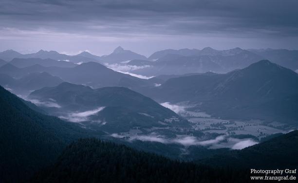 A breathtaking landscape is depicted in this image, showcasing majestic mountains towering into the sky. The scene is enveloped in a blanket of thick clouds, adding an ethereal and mystical quality to the view. The dominant colors of black and a hint of dark blue create a dramatic and moody atmosphere. The foreground features a vast expanse of land, adding depth and perspective to the composition. This serene and awe-inspiring nature scene captures the beauty and grandeur of mountain ranges, ev…