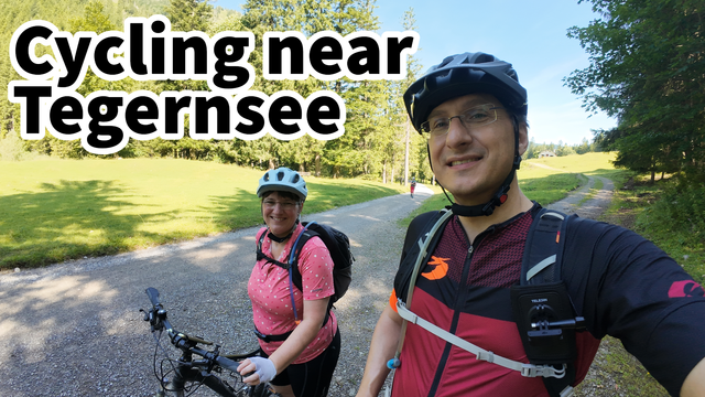 A man and a woman are enjoying a leisurely bike ride together. The man is wearing a helmet and cycling gear, while the woman has a backpack on. The couple is smiling as they pedal along, with the man holding onto the handlebars of their grey bicycle. In the background, there is a tall tree and lush green grass. The man is also wearing glasses, and there is a close-up of his chest. Overall, this image captures a happy and active outdoor moment of cycling.