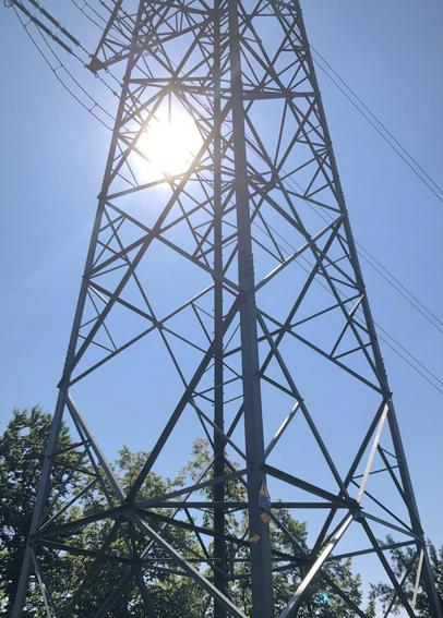 Der mittlere Teil eines Strommastes durch den die Sonne scheint, die Sonne perspektivisch an einer Stelle, wo sich Isolatoren und Stromkabel befinden.