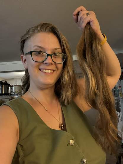 Geneva holds up two feet of their old chopped-off hair next to their new shoulder length hair with a buzzed undercut exposed on one side, splitting their face with a grin. They sport nerdy green glasses, a green denim vest, and a golden mushroom necklace. 
