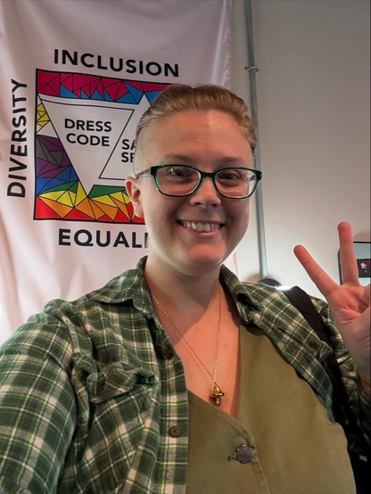 Geneva stands smiling in front of a tapestry showing rainbow and trans color fractals, the words “Inclusion, Diversity, Equality” on the borders, and two nestled triangles saying “Dress Code” and “Safe Space.” Their new haircut is pulled back into a Viking-style braid, revealing both sides of their new undercut. They sport nerdy green glasses, a green denim vest, their dad’s green plaid flannel, and a golden mushroom necklace. 