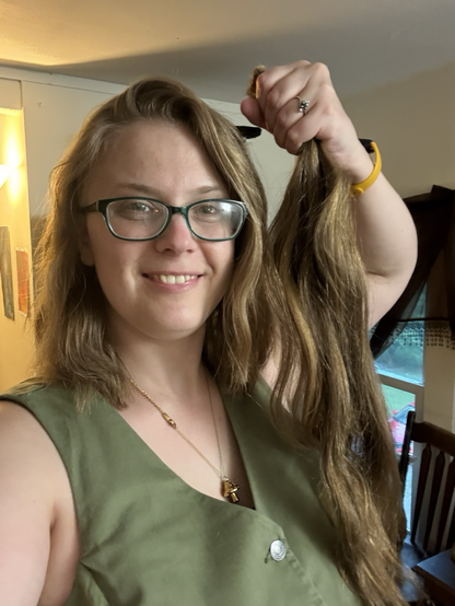 Geneva holds up two feet of their old chopped-off hair next to their new shoulder length hair parted in the middle to cover their undercut, smiling with pride. They sport nerdy green glasses, a green denim vest, and a golden mushroom necklace. 