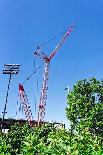 A crane with a two-part boom and a counter-boom and two more structures that the cable that holds up the boom is routed through,  there is a hedge and a tree in front and a bunch of sports lights,  behind it is the fieldhouse structure and behind all that a perfectly clear blue sky.