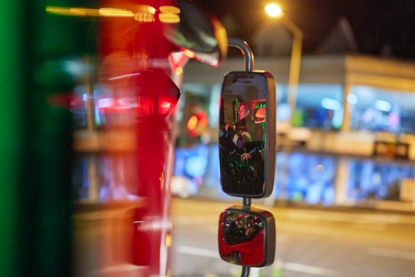 Das Foto zeigt eine Nahaufnahme des Außenspiegels eines großen roten Feuerwehrfahrzeugs. Der Blickwinkel ist von der Seite des Fahrzeugs, leicht nach hinten gerichtet, und das Fahrzeug selbst ist im linken Drittel des Bildes verschwommen und rot leuchtend zu sehen, was auf Bewegung und die aktivierten Warnleuchten hindeutet. Die Oberfläche des Fahrzeugs spiegelt grüne und gelbe Lichter der Ampel wider. Der Hauptfokus liegt auf dem Außenspiegel in der Mitte des Bildes, der klar und scharf ist. Im oberen, größeren Teil des Spiegels ist das verzerrte, aber erkennbare Spiegelbild eines Fahrers zu sehen, der einen Helm mit Visier trägt. Seine Augen sind auf die Straße gerichtet. Darunter, im kleineren Konvexspiegel, ist ein weiteres, noch kleineres und unschärferes Spiegelbild zu erkennen, das ebenfalls Teile des Fahrgastraums  widerspiegelt. Der Hintergrund ist unscharf, aber man kann schemenhaft weitere Gebäude, Lichter und eine Straße bei Nacht erkennen. Die Lichter im Hintergrund sind meist blau, weiß und gelb, was auf eine städtische Umgebung hindeutet. Die gesamte Szene vermittelt den Eindruck von Bewegung und Dringlichkeit in der Dunkelheit. 