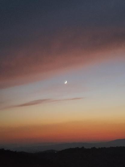 Picture of a sunset with a gibbous moon on the top center and a hilly landscape at the bottom