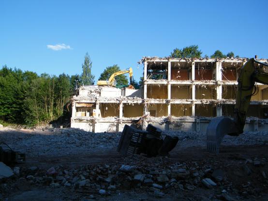 Auf dem Bild sieht man die Reste einen 4 Etagigen Industriegebäudes bei dem schon ein Großteil der Struktur fehlt. Es wirkt wie eine Art riesiger Setzkasten. Auf der linken Seite steht ein Bagger scheinbar auf dem Gebäudeteil welcher schon auf die 2 Etage abgetragen wurde. Er steht aber tatsächlich dahinter, da die Straße höher verläuft. Im Vordergrund es noch eine plane Fläche mit Bauschutt vom davor liegenden Gebäude zu sehen.