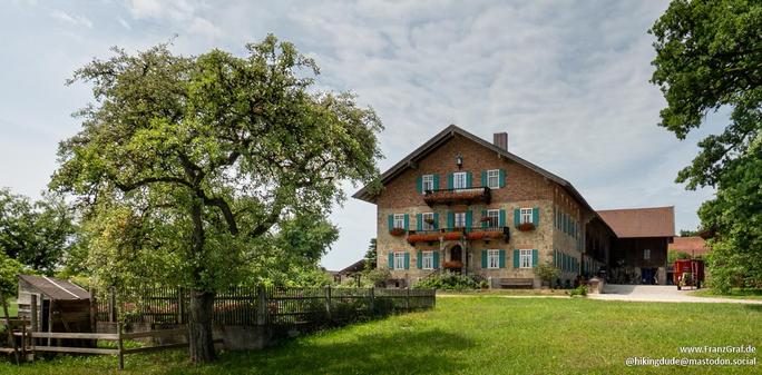 A charming scene of a white house with green shutters, nestled in a rural area. The house is surrounded by a wooden fence, with a lush tree standing tall in the yard. The sky above is a vibrant blue with fluffy white clouds. The grass is a vivid green, creating a picturesque landscape. In the background, another tree can be seen, adding to the natural beauty of the setting. This peaceful and idyllic scene evokes a sense of tranquility and rural charm, making it a perfect retreat from the hustle…