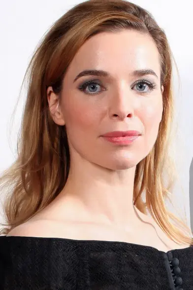 The image features a woman with light brown hair styled in loose waves, parted in the middle, cascading over her shoulders. She has fair skin and striking blue eyes, accentuated by subtle makeup, including mascara and a light pink lipstick. Her expression is neutral, with a soft smile. She is wearing a black off-the-shoulder top with a textured pattern and a row of small black buttons down the front. The background is plain and light-colored, providing a neutral backdrop that highlights her fea…