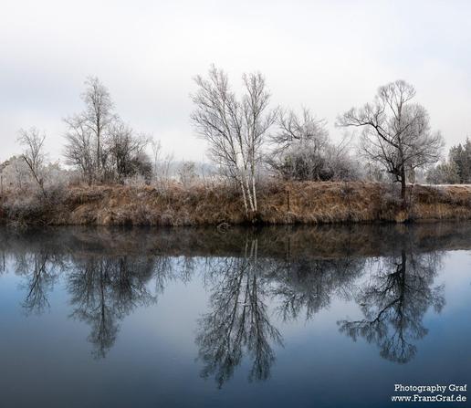 A serene winter landscape is depicted in this image, featuring a body of water surrounded by trees and grass. The photo captures the peaceful reflection of the bare trees in the calm water. The dominant colors in the scene are white and grey, creating a tranquil and cold atmosphere. Various trees of different sizes are scattered throughout the landscape, some with snow on them, adding to the winter aesthetic. The image evokes a sense of tranquility and natural beauty, with the clear sky above a…