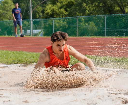 A man with short hair in an orange tank top is crouching in a sand pit while his forward motion knocks up a cloud of sand,  behind him a man is walking on the track with a blue shirt.
