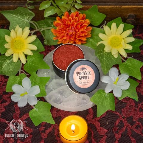 A small round black metal tin of Phoenix Spirit Self-Lighting Incense Tin is displayed on a quartz disc. It contains a red-orange powder, and the label on the lid reads, "Phoenix Spirit / Self-Lighting Incense." This is an exclusive product from Inked Goddess Creations.