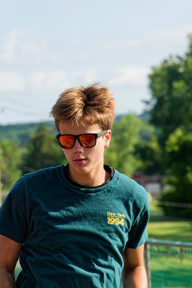A man with medium-short hair has yellow shades and is seen half-torso an up with a bluish shirt that reads New York 1994 in small yellow print with trees in the background