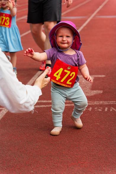 A toddler wearing a purple-pink hat and purple T-shirt has a bib that says 472 in yellow on a red background and light blue pants and little tiny shoes has her back foot off the ground as she advances towards an outstretched hand that can be seen in the background,  the incomplete image of another toddler and adult following them can be seen in the lane to the left
