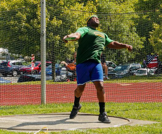 A large black man is caught for a moment while he throws the discus,  seen in his right (your left) hand with his left foot off the ground and shadow visible on a circular platform and his left hand out.  He's looking up and to the right and has blue shorts,  a green shirt that says SYRACUSE CHARGERS and a green and yellow stripe headband